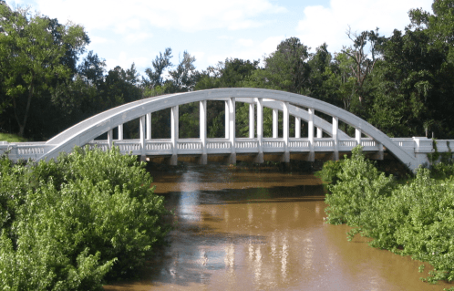 A white arch bridge spans a river, surrounded by lush greenery and trees under a blue sky with scattered clouds.