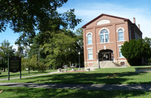 A brick building with large windows, labeled "Johnston Public Library," surrounded by trees and green grass.