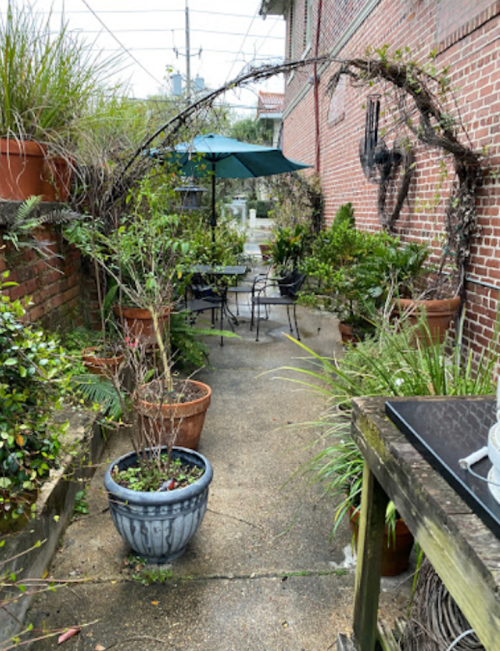 A cozy garden patio with potted plants, a table and chairs, and an umbrella, surrounded by brick walls.