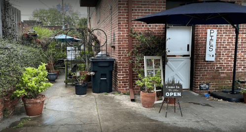 A cozy outdoor space with potted plants, a trash bin, and a sign reading "Come In, We're Open" near a pie shop.