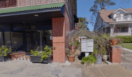 A brick building with a green awning, surrounded by potted plants and a mailbox, on a sunny day.