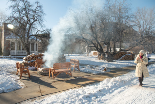 A woman in a winter coat walks past a snowy area with smoke rising from a fire pit and wooden benches nearby.