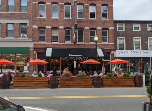 Outdoor dining area of a restaurant with orange umbrellas, surrounded by wooden planters, on a busy street.