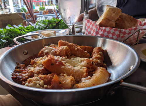 A metal dish filled with pasta in marinara sauce, topped with cheese, served at a restaurant with drinks in the background.