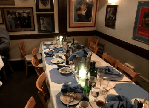 A dining table set with blue napkins, empty plates, and various bottles, surrounded by framed photos on the walls.