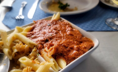 A bowl of pasta with a rich red sauce, served alongside another pasta dish on a blue tablecloth.