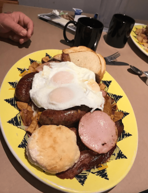 A colorful plate of breakfast featuring eggs, sausage, ham, bacon, a biscuit, and toast, with coffee mugs in the background.