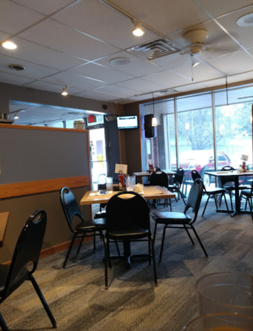 A cozy restaurant interior with empty tables and chairs, large windows, and a TV mounted on the wall.