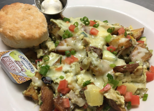 A plate of loaded breakfast nachos with cheese, diced tomatoes, green onions, a biscuit, and a side of syrup.