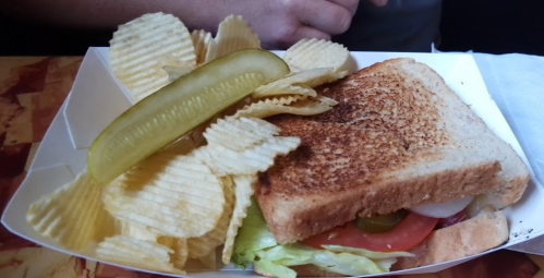 A toasted sandwich with lettuce, tomato, and pickles, served with a side of potato chips on a white tray.