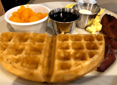 A plate with a waffle, scrambled eggs, bacon, mandarin oranges, and two small cups of syrup and cream.