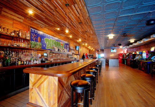 A spacious bar with wooden decor, a long counter, shelves of liquor, and arcade games in a vibrant atmosphere.