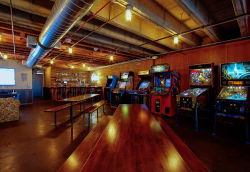 A dimly lit arcade with vintage pinball machines and a long wooden table in a rustic setting.