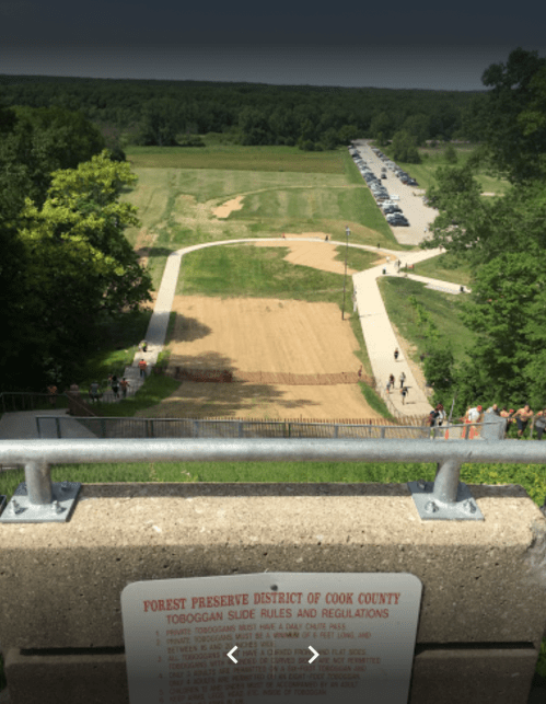 View from a hilltop overlooking a toboggan slide, grassy area, and a parking lot with trees in the background.