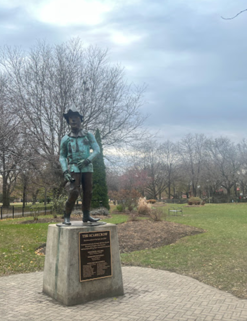 A bronze statue of a cowboy stands on a pedestal in a park, surrounded by bare trees and cloudy skies.