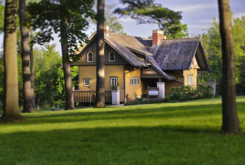 A charming yellow house surrounded by tall trees and lush green grass under a clear sky.