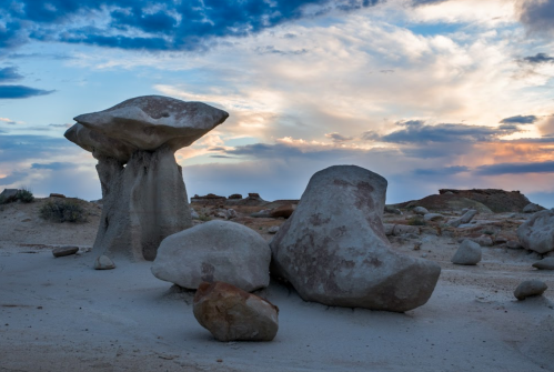 A unique rock formation resembling a mushroom stands amidst scattered boulders under a colorful sky at dusk.