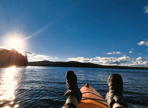 A person's feet in water shoes resting on the edge of an orange kayak, with a scenic lake and sunlit sky in the background.