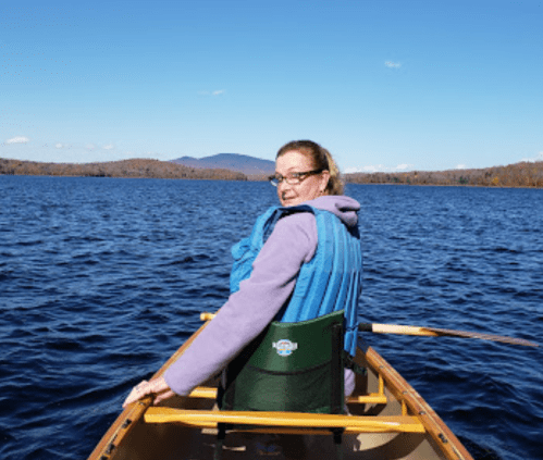 A person in a blue life jacket smiles while rowing a canoe on a calm lake surrounded by autumn-colored trees.
