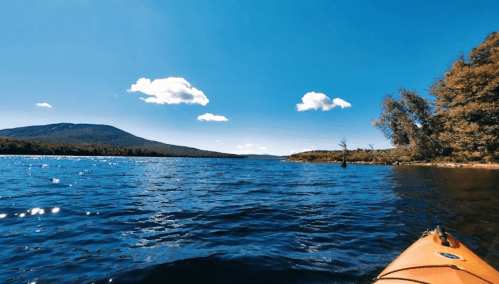 A serene lake view with mountains in the background, bright blue sky, and fluffy clouds, seen from a kayak.