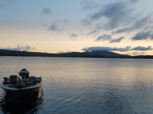 A calm lake at sunset with a small boat floating peacefully in the foreground and mountains in the distance.