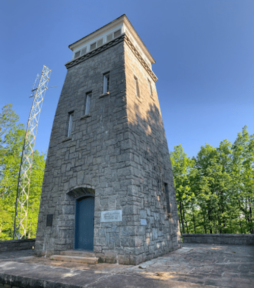 Stone tower with a flat roof, surrounded by trees and a communication tower in the background under a clear blue sky.