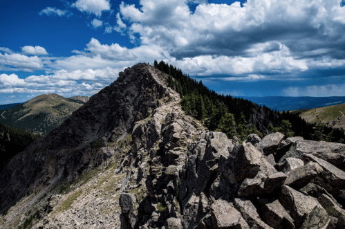 A rocky mountain ridge under a blue sky with scattered clouds, surrounded by lush green forests and distant peaks.