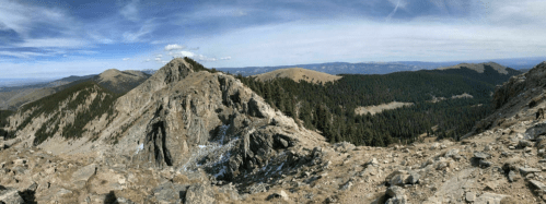 A panoramic view of rugged mountains and valleys under a clear blue sky, showcasing rocky terrain and distant peaks.