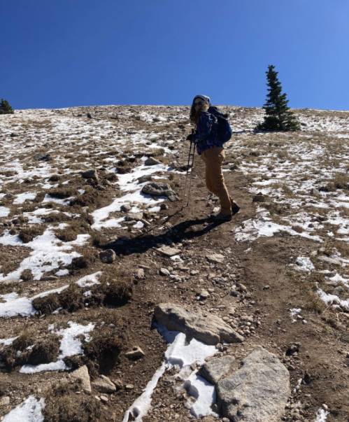 A hiker with trekking poles ascends a snowy, rocky trail under a clear blue sky.
