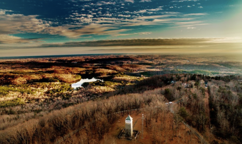 Aerial view of a landscape with rolling hills, a tower, and a lake under a colorful sky at sunset.