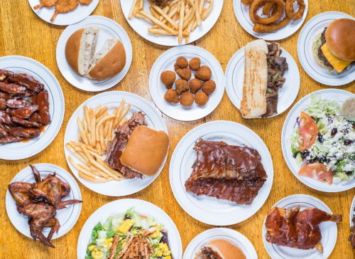 A variety of dishes on plates, including wings, ribs, burgers, fries, salads, and onion rings on a wooden table.