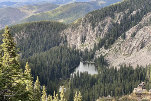 A serene mountain landscape featuring a small lake surrounded by dense evergreen forests and rugged terrain.