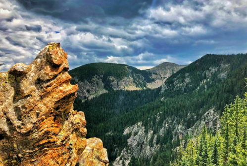 A rocky outcrop overlooks a lush green valley surrounded by mountains under a dramatic cloudy sky.