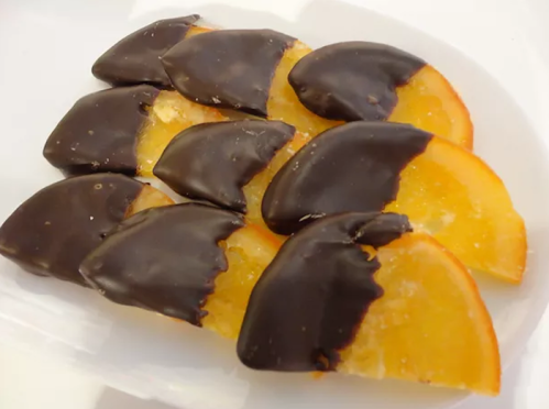 Sliced orange segments partially dipped in dark chocolate, arranged on a white plate.