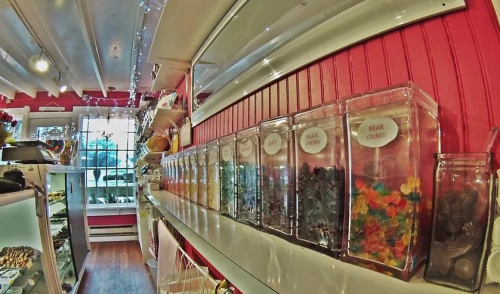 Colorful candy jars line a white shelf in a bright, cheerful candy shop with pink walls and festive lighting.