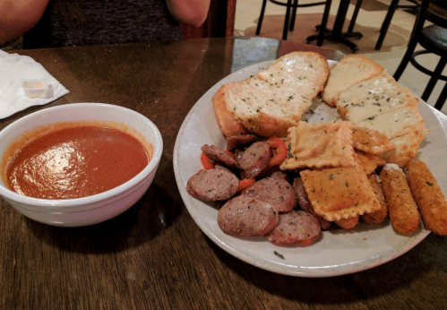 A plate with garlic bread, ravioli, sausage, and mozzarella sticks, alongside a bowl of marinara sauce.