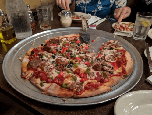 A large pizza on a metal tray, topped with meat, vegetables, and cheese, with slices cut and ready to serve.