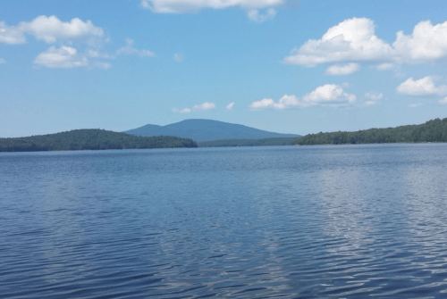 A serene lake with calm waters, surrounded by green hills and a distant mountain under a blue sky with fluffy clouds.