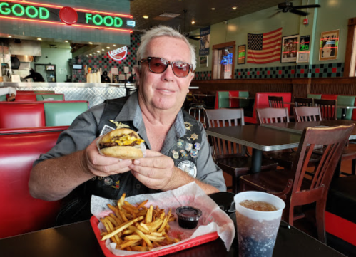 A man in sunglasses holds a burger with fries and a drink in a diner with a retro vibe.
