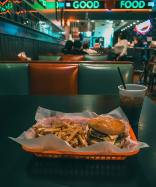 A burger and fries in a basket on a table, with a drink beside it, in a colorful diner setting.