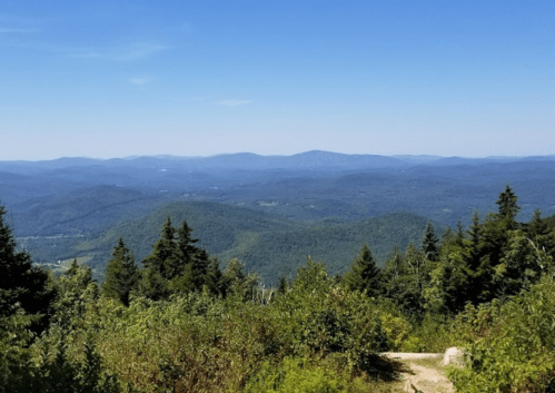 A panoramic view of rolling green mountains under a clear blue sky, with a path leading through lush vegetation.