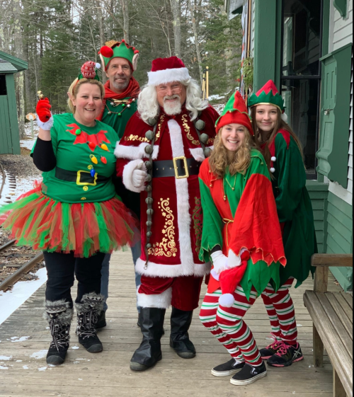 A festive group in holiday attire poses outside, featuring Santa and cheerful elves in a snowy setting.