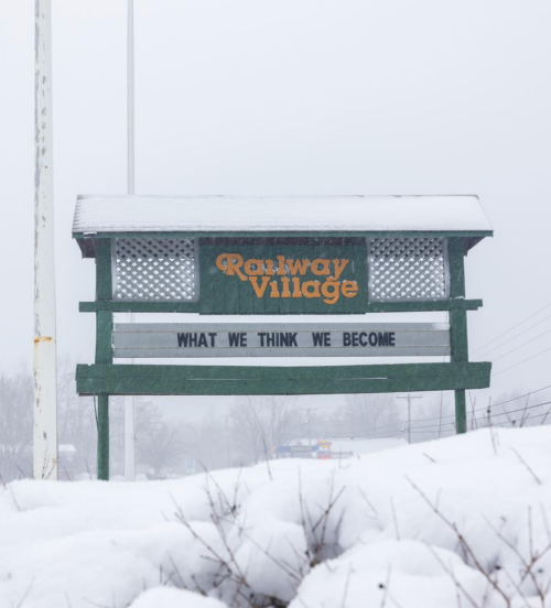 Sign for Railway Village partially covered in snow, featuring the phrase "WHAT WE THINK WE BECOME."