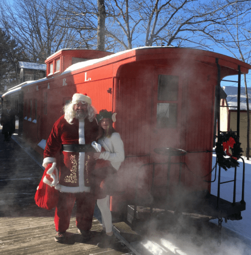 Santa Claus and a woman in festive attire stand outside a red train car, surrounded by snow and steam.