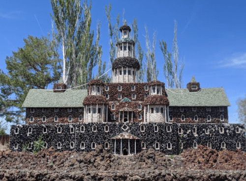 A large, intricately designed building made of stones and wood, featuring multiple towers and a green roof against a blue sky.