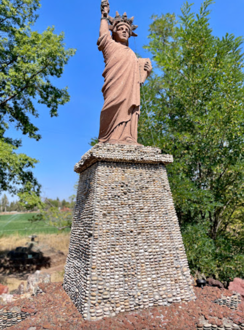 A statue resembling the Statue of Liberty, made from stones, stands on a rocky pedestal surrounded by greenery.