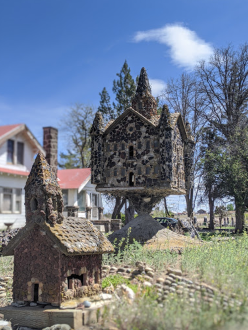 Two whimsical stone houses, one elevated on a mound, surrounded by greenery and trees under a blue sky.