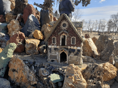 A small decorative house made of stones and shells, surrounded by colorful rocks in a natural setting.