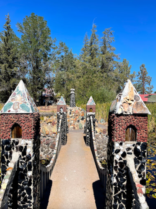 A stone bridge with decorative towers leads into a lush garden under a clear blue sky.