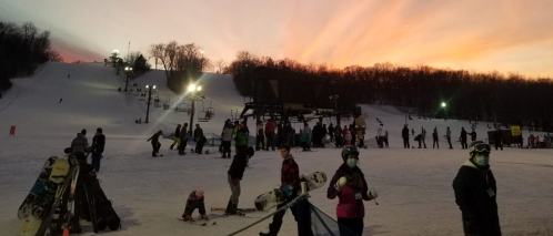 Skiers and snowboarders on a snowy slope at sunset, with a ski lift and trees in the background.